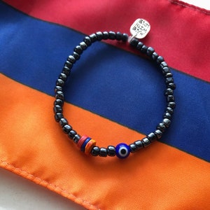 May include: Black beaded bracelet with a blue evil eye bead and colorful accents. The bracelet includes a small silver tag that says "MADE WITH LOVE". The bracelet is displayed on a flag with red, blue, and orange stripes.
