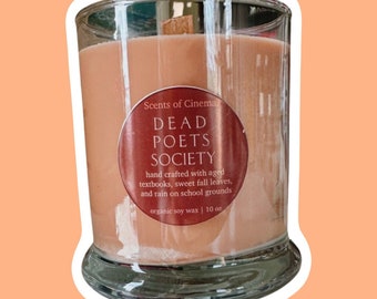 May include: A scented candle in a clear glass jar with a wooden wick. The candle is peach-colored and has a label that reads "DEAD POETS SOCIETY" and "Scents of Cinema." The label also mentions organic soy wax and 10 oz.