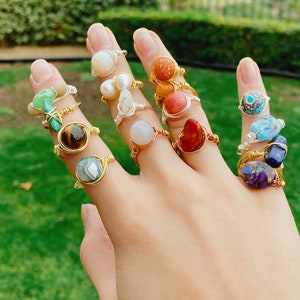 May include: A hand displays a collection of gemstone rings. The rings feature various stones, including tiger's eye, turquoise, and pearls, set in gold, silver, and copper wire. The rings are of different sizes and shapes.