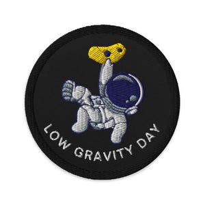 May include: A black embroidered patch with a white astronaut floating in space holding a yellow rock. The text "LOW GRAVITY DAY" is written in white around the astronaut.