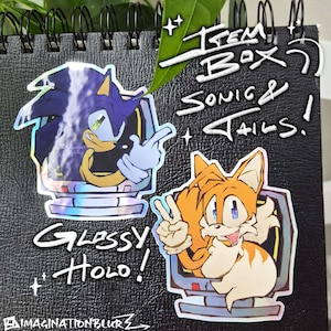 May include: Two glossy holographic stickers featuring Sonic the Hedgehog and Tails from the Sonic the Hedgehog video game series. The stickers are in a computer-like frame with the text "Item Box" and "Sonic & Tails!" written above them.