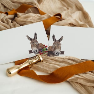 May include: Two white blank cards with donkey illustrations, a gold wax seal, and a brown ribbon on a beige fabric background. The donkey illustrations feature a brown donkey head with flowers.