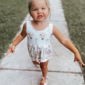 May include: A young girl wearing a white sleeveless dress with a brown cow print and floral accents. She is walking on a sidewalk with green grass on either side.