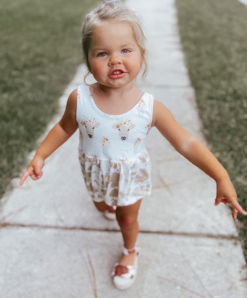 May include: A young girl wearing a white sleeveless dress with a brown cow print and floral accents. She is walking on a sidewalk with green grass on either side.