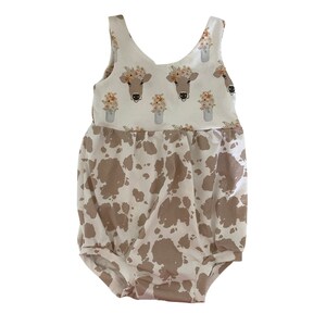 May include: A white and brown baby onesie with a cow print pattern. The top half of the onesie features a floral print with cow faces in mason jars.