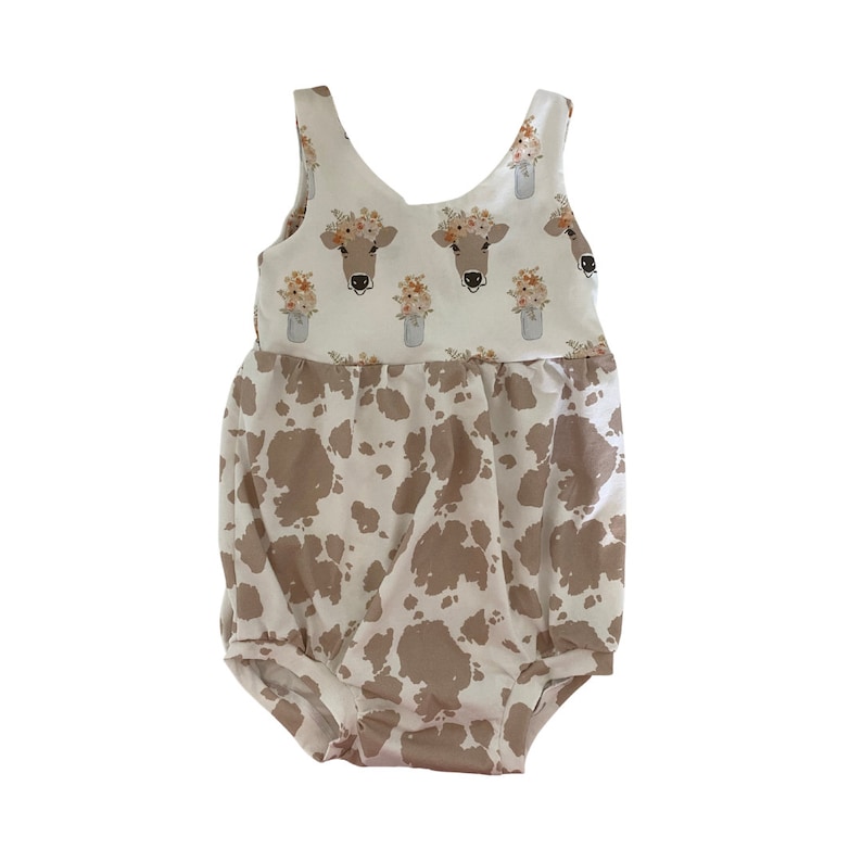 May include: A white and brown baby onesie with a cow print design. The top half of the onesie features a floral print with cow faces in mason jars.