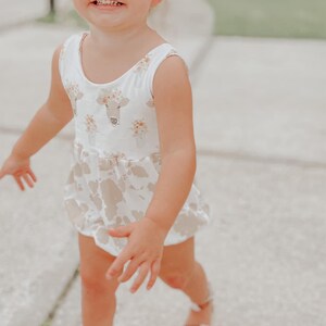 May include: A young child wearing a white and brown sleeveless dress with a cow print and floral design. The child is smiling and walking on a sidewalk.