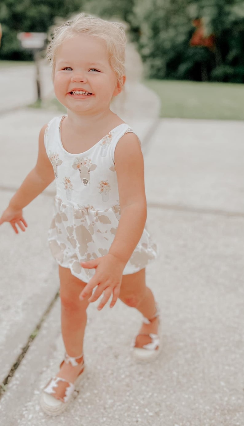 May include: A young child wearing a white and brown sleeveless dress with a cow print and floral design. The child is smiling and walking on a sidewalk.