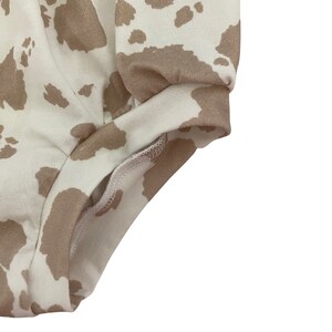May include: Close-up of the bottom edge of a pair of white and brown cow print shorts. The shorts are made of a soft, stretchy fabric and have a comfortable fit.