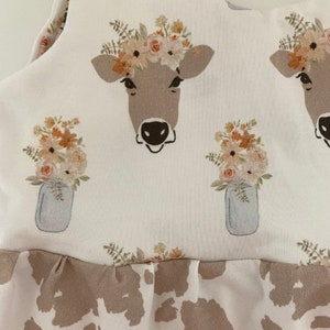 May include: A white sleeveless dress with a brown cow print pattern. The dress has a ruffled hem and features a floral print with a cow wearing a flower crown.