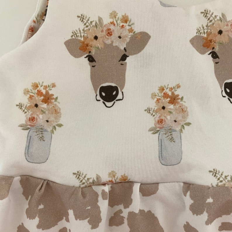 May include: A white sleeveless dress with a brown cow print pattern. The dress has a ruffled hem and features a floral print with a cow wearing a flower crown.