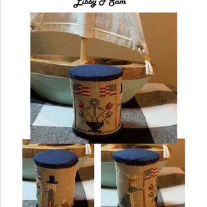 May include: Three different views of a small, cylindrical, fabric-covered box with a blue top. The box is decorated with a cross-stitch design featuring a patriotic theme with red, white, and blue colours. The design includes a small house, a flag, and a flower.