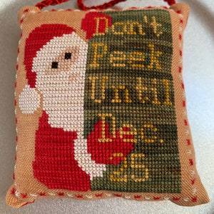 Może przedstawiać: Poduszka w kolorze brązowym i beżowym z czerwono-białym Świętym Mikołajem po lewej stronie i zielono-złotym napisem "Don't Peek Until Dec. 25" po prawej stronie.