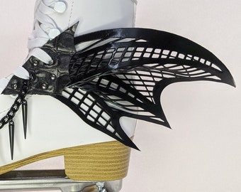Könnte beinhalten: Weißer Rollschuh mit schwarzen Flügeln und silbernen Akzenten. Der Rollschuh hat eine schwarze Kette mit Stacheln, die daran hängen. Die Räder sind braun und weiß.