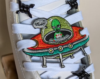Peut inclure: Un patin à roulettes blanc avec un protège-orteils vert et un motif de crâne. Le patin a des lacets blancs avec des breloques de lacets en fil de fer barbelé noir. Un extraterrestre vert dans un patch de soucoupe volante se trouve sur la partie supérieure du patin.