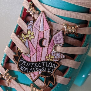 May include: A teal roller skate with pink laces and decorative gold-colored accents. The skate features a patch with a crystal design and the text "PROTECTION FROM ASSHOLES". The toe stop has gold spikes and a ring.
