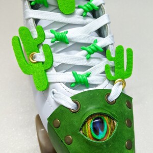 Puede incluir: Patín de ruedas blanco con abalorios en forma de cactus verdes en los cordones y una puntera de cuero verde con un diseño de ojo de pluma de pavo real.
