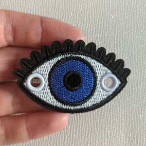 May include: A blue evil eye patch with white trim and black eyelashes. The patch has two white grommets on either side.