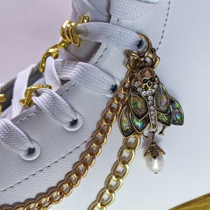 May include: White roller skate with gold chain and a decorative charm. The charm is a skull-shaped insect with iridescent wings and a dangling pearl. The skate has white laces and gold accents.