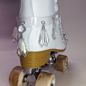 May include: White roller skates with a clear plastic strap and crystal embellishments. The skates have brown wheels and a white leather boot.
