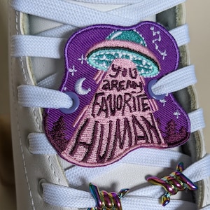 Op de afbeelding: Witte rolsschaats met een paarse en roze alien-patch met de tekst "You are my favorite human" en regenboogkleurige prikkeldraad-veters.
