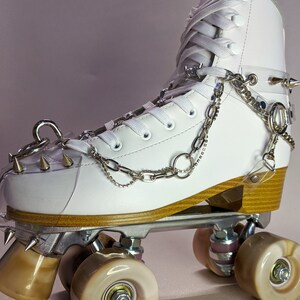 May include: White roller skate with brown wheels and silver chains and spikes. The skate is laced up and has a white leather upper.