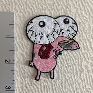 Puede incluir: Personaje de dibujos animados rosa con ojos grandes blancos y pupilas negras. Los ojos tienen venas negras y el personaje tiene una boca roja y una nariz gris. El personaje mide aproximadamente 5 cm de alto.