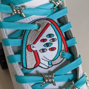 Puede incluir: Patín blanco con cordones turquesa y abalorios de alambre de púas plateado. Un parche bordado de colores con una cara de dibujos animados y varios ojos está unido a los cordones.