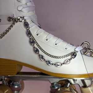 Könnte beinhalten: Weiße Rollschuhe mit braunen Rollen und silbernen Ketten, die an der Stiefelette befestigt sind. Die Rollschuhe stehen auf einer hellrosa Oberfläche.