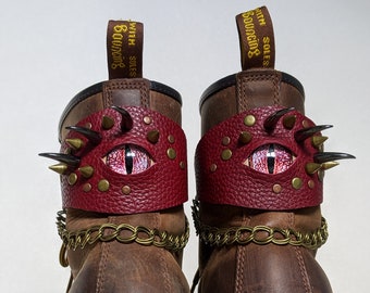 Puede incluir: Botas de cuero marrón con un brazalete de cuero rojo en forma de ojo de dragón con tachuelas doradas y una cadena dorada. El brazalete tiene un ojo rojo con un iris dorado y una pupila negra. Las botas tienen el texto "with bouncing soles" en la parte posterior.
