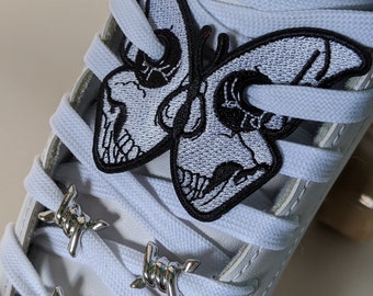 Könnte beinhalten: Weiße Sneaker mit schwarzen Schmetterlings- und silbernen Stacheldraht-Schnallen. Der Schmetterling hat ein Totenkopfdesign und ist auf die Schnürsenkel gestickt.