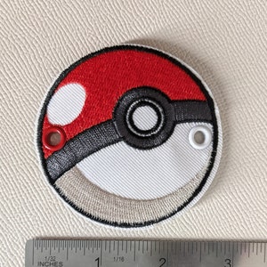 Puede incluir: Un parche bordado rojo, blanco y negro de una Poké Ball, una bola redonda con la parte superior roja, un centro blanco y una banda negra alrededor del centro. El parche tiene dos ojales blancos en el lateral.