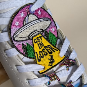 Peut inclure: Patin à roulettes en cuir blanc avec un patch UFO rose et vert et un patch jaune qui dit "Get in Loser" sur les lacets. Les lacets ont également des fermoirs à lacets en fil de fer barbelé métalliques irisés.