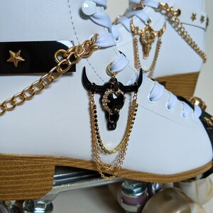 May include: White roller skates with gold and black decorative accents. The skates feature gold chain detailing, black bull skull charms, and a black strap with a gold star. The wheels are a light brown colour.