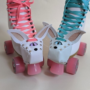 May include: A pair of white roller skates with pink and teal laces. Each skate has a white leather bunny face attached, complete with ears, eyes, and nose. The wheels are pink.