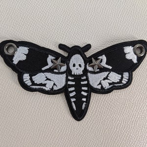 May include: Black and white embroidered moth patch with a skull and star accents. The moth design features a skeleton-like pattern on the wings and body. The patch has two metal grommets and is suitable for clothing or accessories.