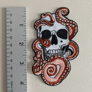 May include: Embroidered patch featuring a white skull with black eyes and teeth, wrapped by a pink and orange octopus tentacle.