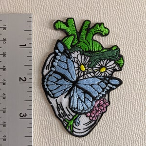 May include: Embroidered patch of a blue butterfly with white and yellow flowers inside a green heart. The heart is surrounded by white fabric.