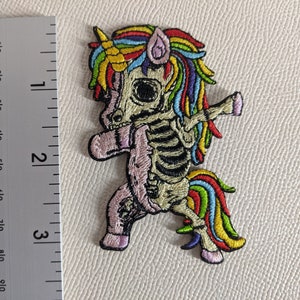 Peut inclure: Un patch brodé coloré représentant une licorne squelette avec une crinière et une queue arc-en-ciel. La licorne est en position de danse, une jambe levée.