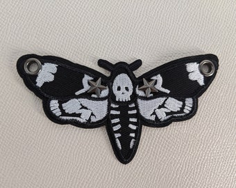 Könnte beinhalten: Schwarz-weißer, gestickter Mottenaufnäher mit Schädel- und Sternakzenten. Das Motten-Design weist ein skelettartiges Muster auf den Flügeln und dem Körper auf. Der Aufnäher hat zwei Metallösen und ist für Kleidung oder Accessoires geeignet.
