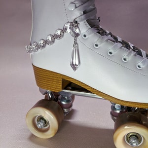 May include: White roller skate with a crystal charm dangling from the laces. The skate has brown wheels and a wooden base.