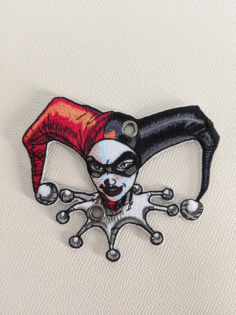 May include: Embroidered patch featuring Harley Quinn, a comic book character, with a red and black jester hat and a white and gray costume. The patch has two metal eyelets.