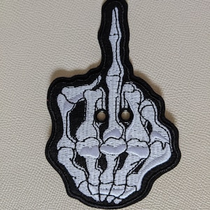 May include: Embroidered patch featuring a white skeleton hand giving the middle finger, outlined in black. The patch has a black border and two eyelets near the top. This patch is suitable for jackets, bags, or other fabric items.