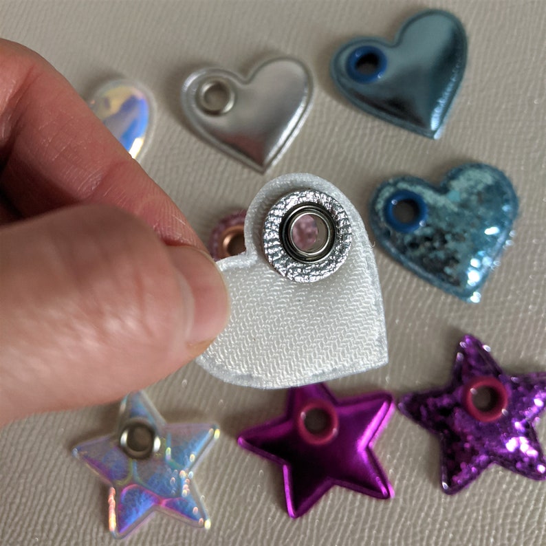 May include: A collection of heart and star-shaped embellishments with eyelets. The embellishments are made of different materials, including iridescent, glitter, and fabric. The embellishments are in various colors, including silver, blue, pink, and purple.