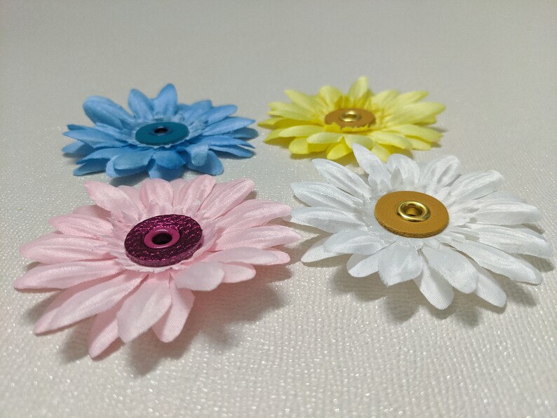 May include: Four fabric flowers with different colored centers. The flowers are light blue, pink, white, and yellow. Each flower has a metal button in the center.