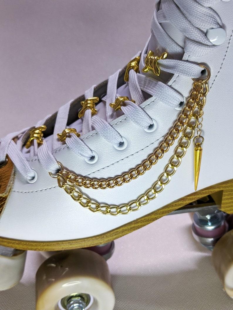 Peut inclure: Gros plan d'un patin &agrave; roulettes blanc avec des d&eacute;tails dor&eacute;s. Le patin comporte des breloques dor&eacute;es sur les lacets et une cha&icirc;ne dor&eacute;e avec un pendentif en forme de pointe. Les roues sont de couleur beige clair, et le patin a une bordure dor&eacute;e.