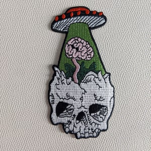 Puede incluir: Un parche bordado que representa una calavera con un cerebro en su interior, siendo abducido por un platillo volante. El cerebro es rosa, el platillo es rojo y blanco, y la calavera es blanca con detalles negros.