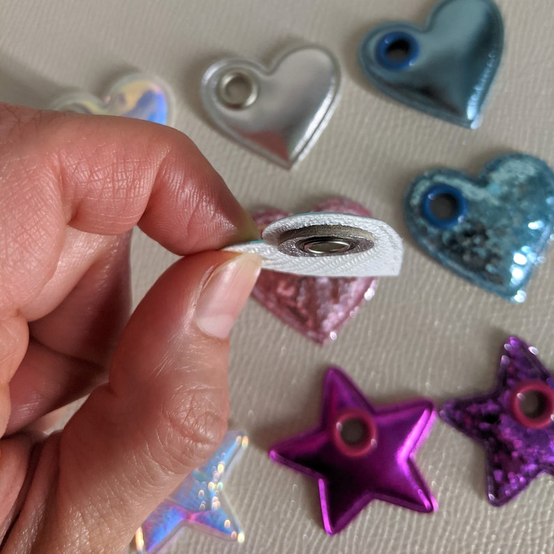 May include: Assortment of colorful heart and star-shaped patches with a metallic finish. The patches have a hole in the center and are designed to be sewn or ironed onto fabric.