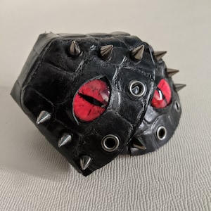 May include: Black leather mask with red eyes and silver studs. The mask has a textured surface and is decorated with spikes.