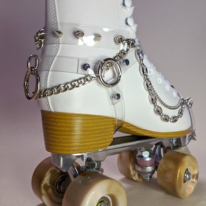 May include: White roller skate with silver metal hardware, brown wheels, and a wooden heel. The skate features clear straps with silver spikes and a chain detail. A skull charm is visible on the side.